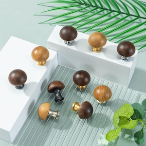 Könnte beinhalten: Eine Sammlung von Holzschrankknöpfen in verschiedenen Braun- und Naturholztönen. Die Knöpfe haben Metallbasen in Silber- und Goldausführung. Einige Knöpfe werden auf einer weißen Plattform und einer transparenten Glasoberfläche präsentiert.