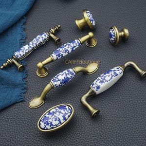 May include: A collection of cabinet hardware featuring brass-toned handles and knobs with white ceramic accents. The ceramic pieces are decorated with a blue floral pattern. The hardware includes various handle styles and knob designs.