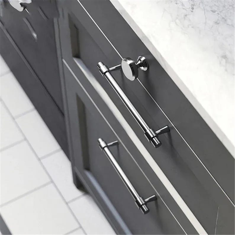 Peut inclure: Gros plan sur une armoire grise &agrave; deux tiroirs, avec des poign&eacute;es de barre chrom&eacute;es polies et un bouton rond. L'armoire est pos&eacute;e contre un comptoir en marbre blanc. Les poign&eacute;es mesurent environ 20 cm de long.