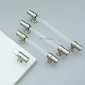 K&ouml;nnte beinhalten: Satz von Schrankgriffen mit modernem Design. Jeder Griff hat einen transparenten, zylindrischen Acrylgriff und Metallbeschl&auml;ge mit geb&uuml;rsteter Nickel-Oberfl&auml;che. Die Griffe werden auf einer hellgrauen Oberfl&auml;che gezeigt, mit dem Markennamen "CARFTBoutique".