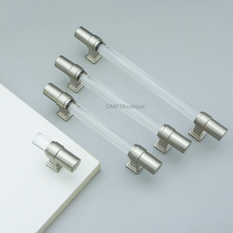 K&ouml;nnte beinhalten: Satz von Schrankgriffen mit modernem Design. Jeder Griff hat einen transparenten, zylindrischen Acrylgriff und Metallbeschl&auml;ge mit geb&uuml;rsteter Nickel-Oberfl&auml;che. Die Griffe werden auf einer hellgrauen Oberfl&auml;che gezeigt, mit dem Markennamen "CARFTBoutique".