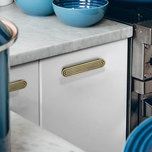 Puede incluir: Primer plano de un armario blanco con dos tiradores de caj&oacute;n horizontales de color lat&oacute;n y forma ovalada. Los tiradores tienen un dise&ntilde;o acanalado. Se ven dos cuencos azules en el fondo, junto con una encimera de m&aacute;rmol.