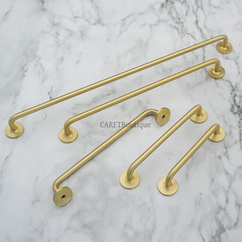 May include: A collection of gold-colored metal towel bars in various sizes. The bars have a simple, curved design with circular mounting plates. The image is taken on a marble surface. The text "CARFTBoutique" is visible.