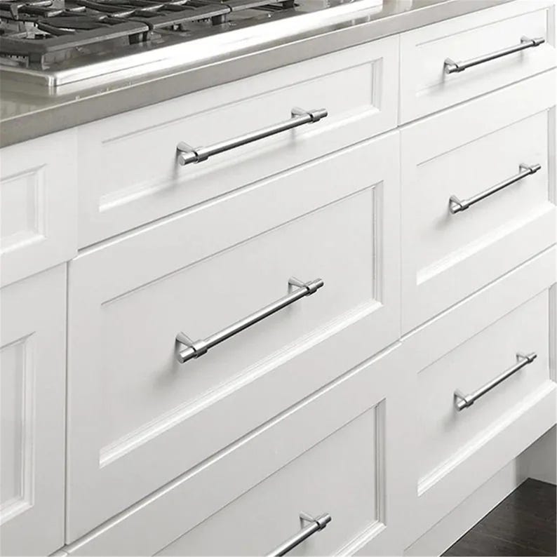 Peut inclure: Gros plan sur des armoires de cuisine blanches avec des poign&eacute;es de barre argent&eacute;es. Les armoires ont une conception de panneaux encastr&eacute;s et sont dispos&eacute;es en une configuration &agrave; plusieurs tiroirs. Les poign&eacute;es sont align&eacute;es horizontalement.