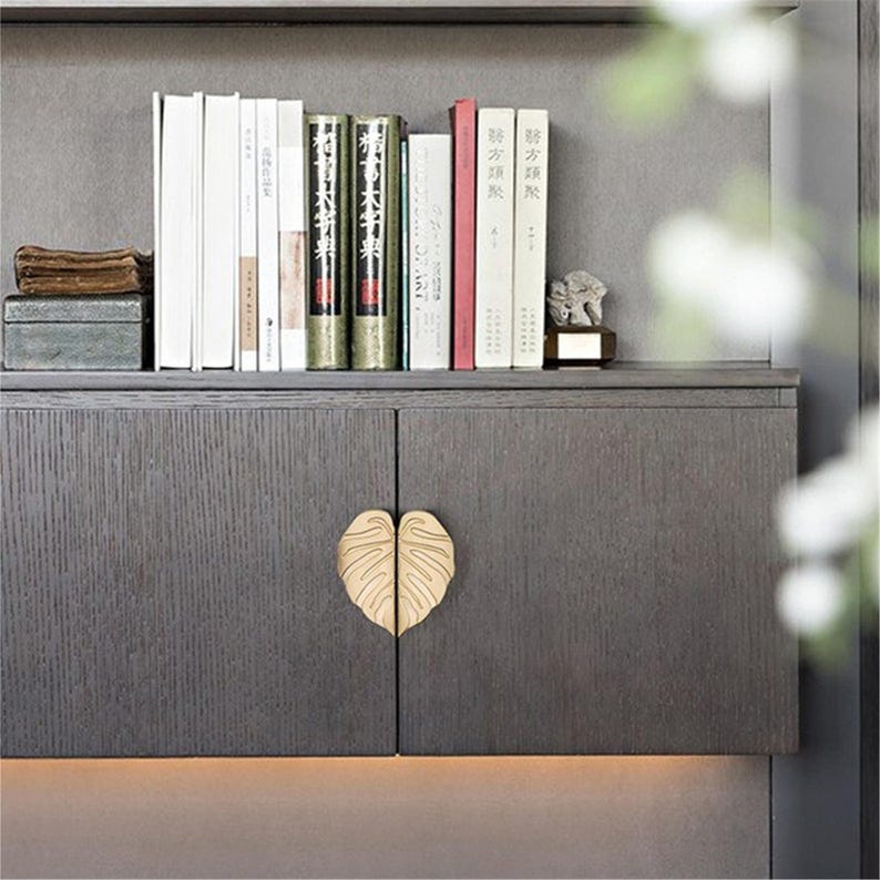 May include: A dark gray cabinet with a leaf-shaped gold handle. The cabinet doors are closed. A shelf above holds books of various sizes and colors, along with a small decorative statue.