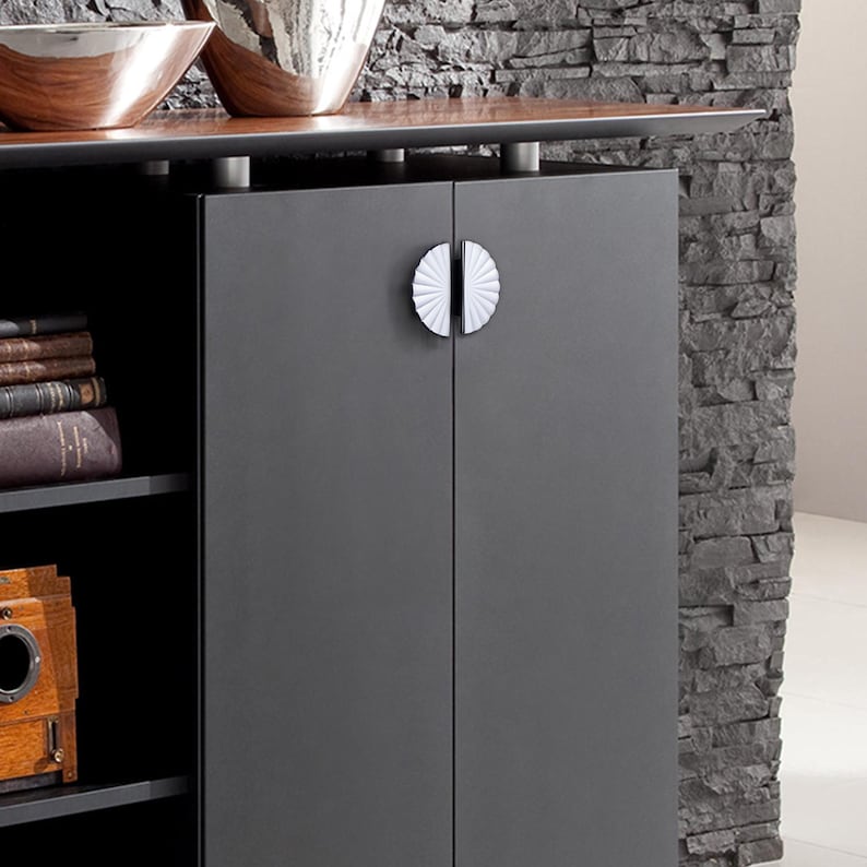 May include: A dark grey cabinet with two doors and white, fan-shaped handles. The cabinet has a wooden top with decorative bowls. Books and a vintage camera are visible on the shelves.