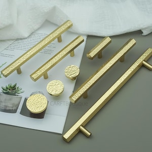 May include: A selection of gold-coloured cabinet hardware, including handles and knobs. The handles have a textured, hammered finish, and the knobs are round with a similar texture. The hardware is displayed on a neutral background. Measurements are in cm.