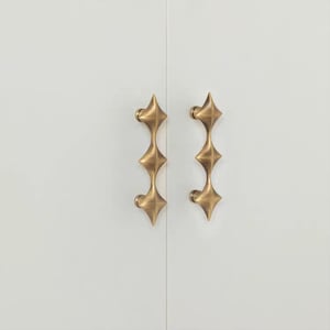 以下が含まれることがあります： 星型のデザインが施された、アンティーク真鍮製のキャビネットプルが2つ。白いキャビネットドアに垂直に取り付けられています。星型のデザインは、プルの長さに沿って3回繰り返されています。