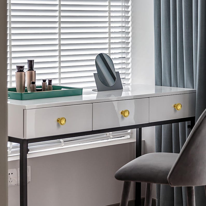 May include: A white vanity table with three drawers, gold knobs, and a black metal frame. A green tray with beauty products sits on the table, along with a small oval mirror. A gray chair is positioned next to the table.