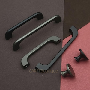 Op de afbeelding: Een assortiment kastbeslag in verschillende tinten grijs en zwart. De collectie bevat verschillende handgrepen en twee knopstijlen. De handgrepen hebben een gebogen ontwerp en de knoppen hebben een afgeronde bovenkant. De achtergrond is verdeeld in bruine en roze secties.