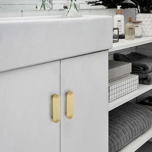 Puede incluir: Un mueble de ba&ntilde;o blanco con dos tiradores dorados de forma ovalada. El mueble forma parte de un dise&ntilde;o de ba&ntilde;o moderno, con estantes que muestran toallas, cajas y art&iacute;culos de tocador. Los tiradores miden unos 10 cm de largo.