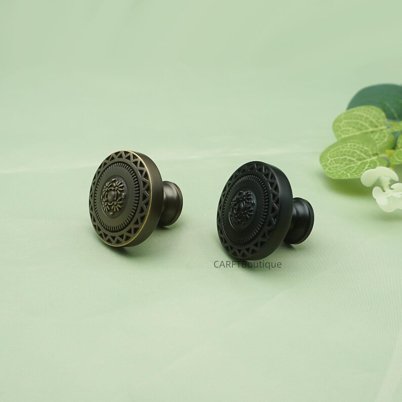 May include: Two decorative cabinet knobs. One is bronze-colored, the other is black. Both have a circular design with a central floral motif and a geometric border. The knobs are likely made of metal and are suitable for furniture or cabinetry.