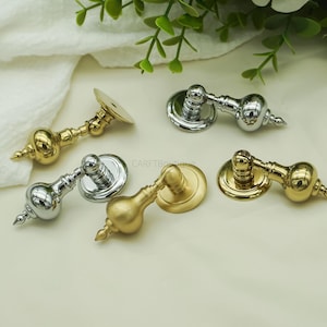 Könnte beinhalten: Eine Auswahl an dekorativen Schrankgriffen in Gold- und Silberausführung. Jeder Griff hat eine runde Basis, einen dekorativen Griff und ein spitzes Ende. Die Griffe sind auf einer weißen Oberfläche mit grünem Laub im Hintergrund angeordnet. Der Text "CARFTBoutique" ist sichtbar.