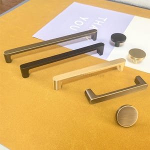 May include: A selection of cabinet hardware in bronze, black, and gold finishes. The image displays bar pulls and round knobs, offering a variety of styles for home improvement. The background includes a paper with the text "THANK YOU".