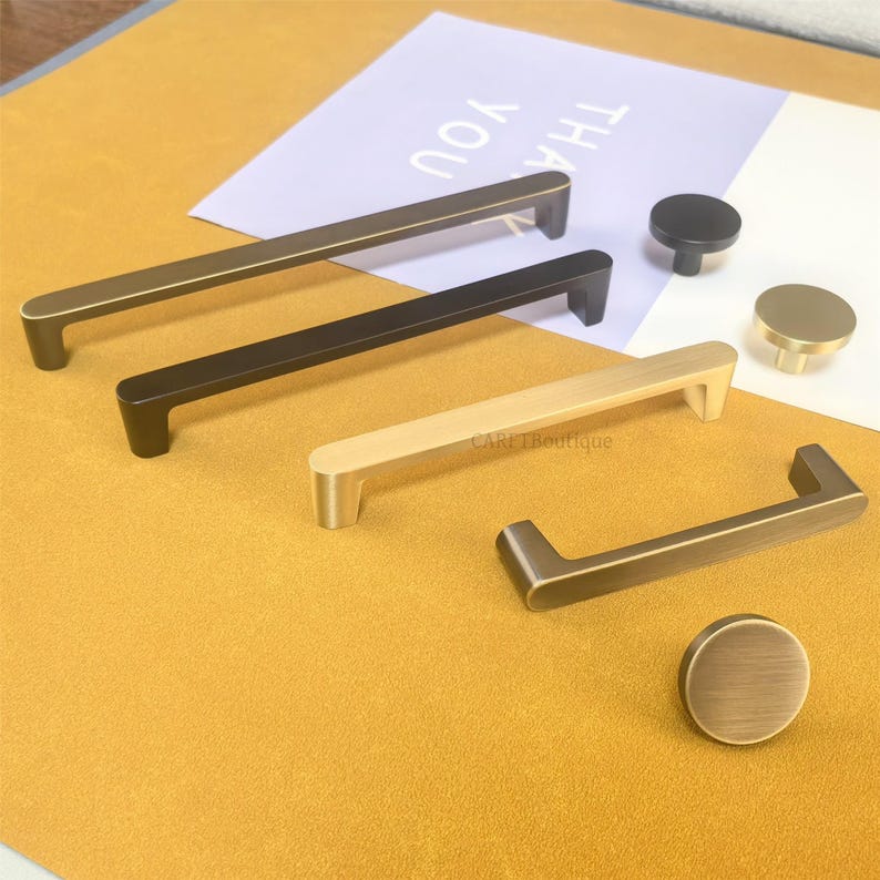 May include: A selection of cabinet hardware in bronze, black, and gold finishes. The image displays bar pulls and round knobs, offering a variety of styles for home improvement. The background includes a paper with the text "THANK YOU".