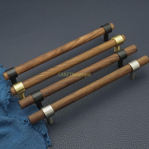 以下が含まれることがあります： 金属アクセントが付いた4つの木製キャビネットプル。プルは、黒、金、銀色の金属製ハードウェアを備えた濃い木目仕上げです。プルは濃い青色の背景に対して斜めに配置されています。「CARETBoutique」の文字が見えます。