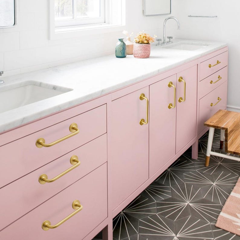 May include: A pink bathroom vanity with gold-colored hardware. The vanity has multiple drawers and cabinet doors. A white countertop with a sink is visible, along with a small wooden stool and patterned floor tiles.