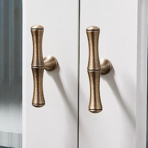 K&ouml;nnte beinhalten: Zwei bronzefarbene Schrankgriffe mit Bambus-Design sind an einer wei&szlig;en Schrankt&uuml;r montiert. Die Griffe haben eine zylindrische Form mit leicht strukturierter Oberfl&auml;che und abgerundeten Enden. Beschl&auml;ge f&uuml;r die Inneneinrichtung.