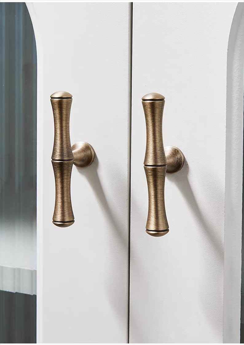 K&ouml;nnte beinhalten: Zwei bronzefarbene Schrankgriffe mit Bambus-Design sind an einer wei&szlig;en Schrankt&uuml;r montiert. Die Griffe haben eine zylindrische Form mit leicht strukturierter Oberfl&auml;che und abgerundeten Enden. Beschl&auml;ge f&uuml;r die Inneneinrichtung.