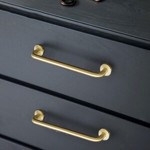 May include: Close-up of a dark grey chest of drawers featuring two brass bar handles. The handles are curved and attached to the drawer fronts with circular mounts. The wood grain is visible.
