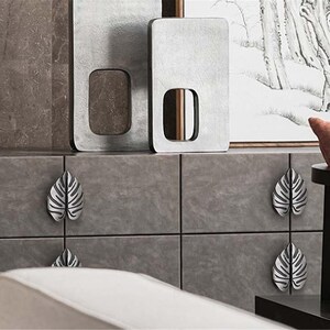 May include: A close-up of a gray cabinet with silver leaf-shaped drawer pulls. Two large, rectangular, gray decorative panels with cut-out shapes are above the cabinet. The cabinet is gray with a white sofa in the foreground.