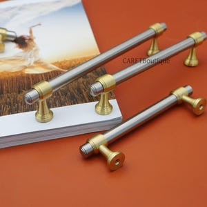 Pode incluir: Ferragens de arm&aacute;rio com acabamento em n&iacute;quel escovado e lat&atilde;o. As al&ccedil;as t&ecirc;m uma barra cil&iacute;ndrica com detalhes em lat&atilde;o em cada extremidade e uma base de lat&atilde;o. O hardware &eacute; exibido em uma superf&iacute;cie branca e um fundo laranja.