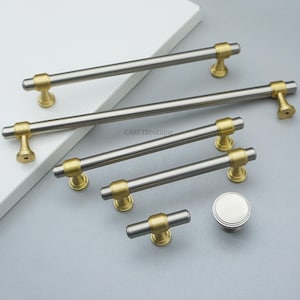 K&ouml;nnte beinhalten: Eine Sammlung von Schrankbeschl&auml;gen, darunter mehrere Stangengriffe in geb&uuml;rstetem Nickel und Gold sowie ein runder Knopf. Die Hardware wird auf einer wei&szlig;en Oberfl&auml;che und einem grauen Hintergrund pr&auml;sentiert. Der Text "CARFTBoutique" ist sichtbar.