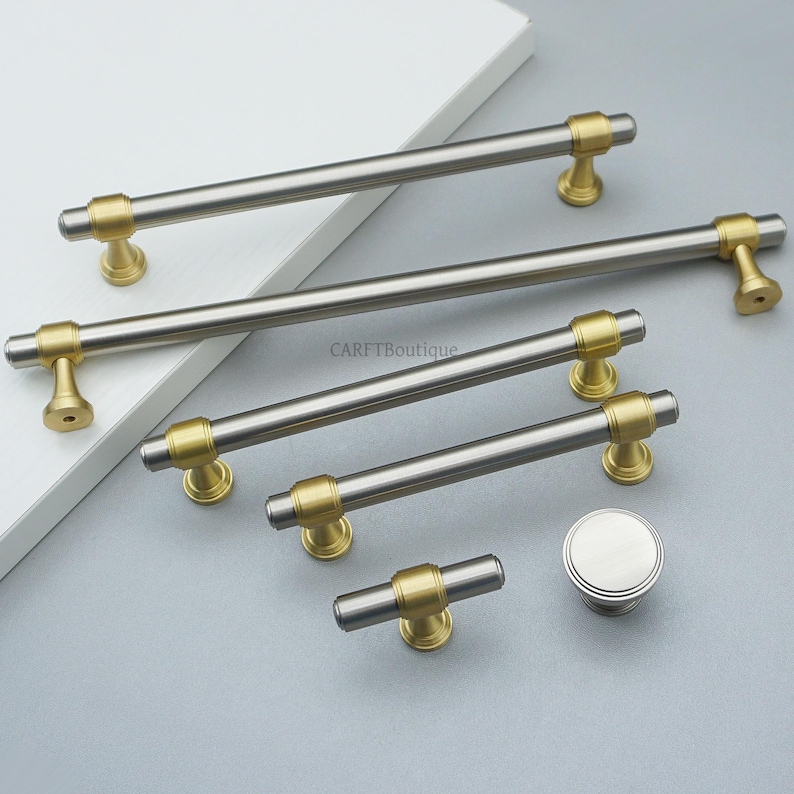 K&ouml;nnte beinhalten: Eine Sammlung von Schrankbeschl&auml;gen, darunter mehrere Stangengriffe in geb&uuml;rstetem Nickel und Gold sowie ein runder Knopf. Die Hardware wird auf einer wei&szlig;en Oberfl&auml;che und einem grauen Hintergrund pr&auml;sentiert. Der Text "CARFTBoutique" ist sichtbar.