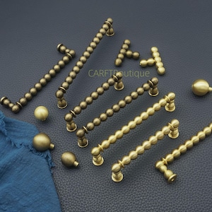 Puede incluir: Una variedad de herrajes de gabinete de latón antiguo, que incluyen manijas y perillas. Las manijas están adornadas con un diseño de cuentas, y las perillas tienen forma redondeada. El hardware se muestra sobre una superficie oscura, con el texto "CARFTboutique".