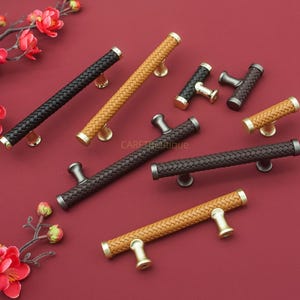 Könnte beinhalten: Eine Sammlung dekorativer Schrankgriffe in verschiedenen Farben, darunter Schwarz, Braun und Beige. Die Griffe haben eine geflochtene, lederartige Textur und gold- oder silberfarbene Beschläge. Maße in Zentimetern.