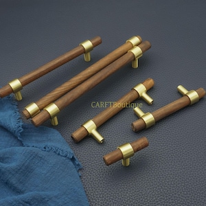 以下が含まれることがあります： 真鍮のアクセントが付いた木製キャビネットプル。プルは円筒形で、濃い木材仕上げで、真鍮のリングとT字型の真鍮ハンドルが付いています。「CARFTBoutique」の文字が見えます。