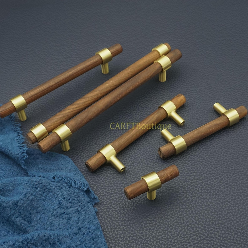 以下が含まれることがあります： 真鍮のアクセントが付いた木製キャビネットプル。プルは円筒形で、濃い木材仕上げで、真鍮のリングとT字型の真鍮ハンドルが付いています。「CARFTBoutique」の文字が見えます。