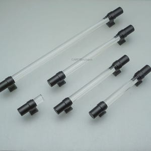 K&ouml;nnte beinhalten: Eine Sammlung von transparenten Acryl-Schrankgriffen mit schwarzen Metallenden und -basen. Die Griffe sind in verschiedenen Gr&ouml;&szlig;en und Formen erh&auml;ltlich, darunter Stangen- und Knaufstile. Die Hardware wird auf einer hellgrauen Oberfl&auml;che pr&auml;sentiert.