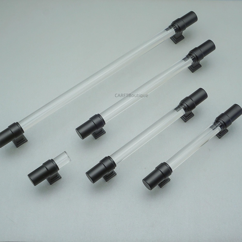 K&ouml;nnte beinhalten: Eine Sammlung von transparenten Acryl-Schrankgriffen mit schwarzen Metallenden und -basen. Die Griffe sind in verschiedenen Gr&ouml;&szlig;en und Formen erh&auml;ltlich, darunter Stangen- und Knaufstile. Die Hardware wird auf einer hellgrauen Oberfl&auml;che pr&auml;sentiert.