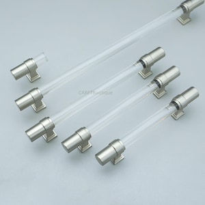 K&ouml;nnte beinhalten: Eine Sammlung von Schrankgriffen aus klarem Acryl und geb&uuml;rstetem Nickel. Die Griffe haben einen zylindrischen Acrylmittelteil und sind auf quadratischen Nickelbasen montiert. Die Hardware ist auf einer hellgrauen Oberfl&auml;che angeordnet. Der Text "CARFTBoutique" ist sichtbar.