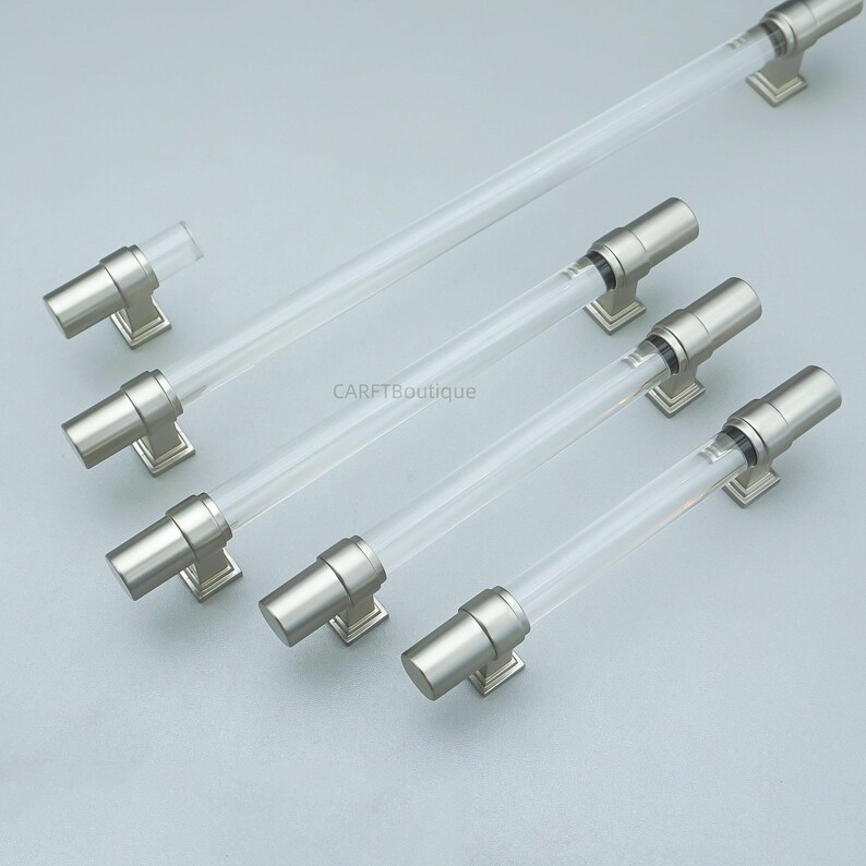 K&ouml;nnte beinhalten: Eine Sammlung von Schrankgriffen aus klarem Acryl und geb&uuml;rstetem Nickel. Die Griffe haben einen zylindrischen Acrylmittelteil und sind auf quadratischen Nickelbasen montiert. Die Hardware ist auf einer hellgrauen Oberfl&auml;che angeordnet. Der Text "CARFTBoutique" ist sichtbar.