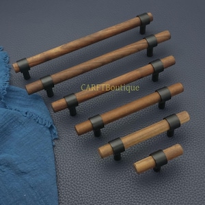 以下が含まれることがあります： 黒い金属のアクセントが付いた木製キャビネットプル。プルは様々な長さで、円筒形の木製ハンドルと中央に黒い金属バンドが付いています。「CARFTBoutique」の文字が表面に見えます。