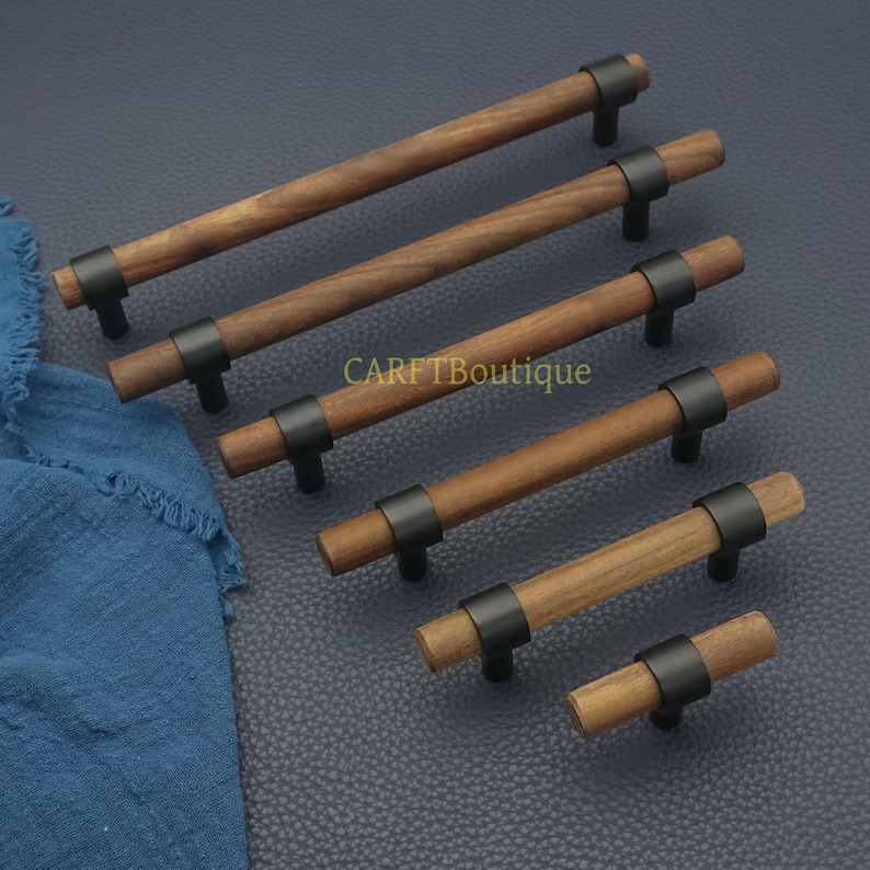 以下が含まれることがあります： 黒い金属のアクセントが付いた木製キャビネットプル。プルは様々な長さで、円筒形の木製ハンドルと中央に黒い金属バンドが付いています。「CARFTBoutique」の文字が表面に見えます。