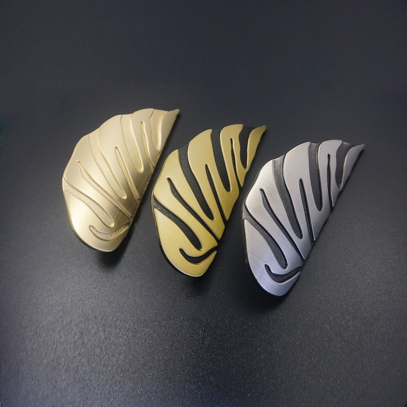 May include: Three decorative metal leaf-shaped cabinet pulls in gold, brass, and silver tones. Each pull features a textured, ridged design, and they are arranged on a dark surface. These hardware pieces add a touch of elegance to furniture.