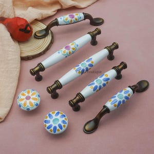May include: A collection of decorative cabinet hardware featuring white ceramic handles and knobs with colorful floral patterns. The handles have a bronze finish and are in various shapes and sizes, including pulls and knobs. The hardware is designed for furniture and home decor.