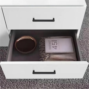 May include: White bedside cabinet with two drawers, one open. The open drawer reveals a brown tray, a book titled "FAHRENHEIT 451", and a folded beige blanket. Black drawer pulls provide contrast.