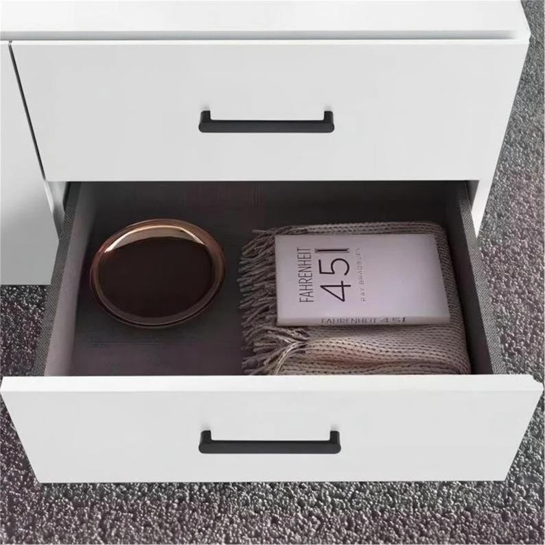 May include: White bedside cabinet with two drawers, one open. The open drawer reveals a brown tray, a book titled "FAHRENHEIT 451", and a folded beige blanket. Black drawer pulls provide contrast.