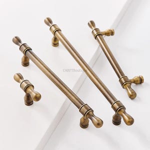 K&ouml;nnte beinhalten: Eine Sammlung antiker Messing-Schrankgriffe und -Kn&ouml;pfe. Die Hardware hat eine zylindrische Stange mit dekorativen Endst&uuml;cken. Die Griffe sind in verschiedenen Gr&ouml;&szlig;en erh&auml;ltlich und haben eine warme, gealterte Oberfl&auml;che. Der Markenname "CARFTBoutique" ist sichtbar.