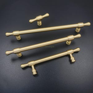 K&ouml;nnte beinhalten: Ein Set aus vier messingfarbenen Schrankgriffen und -kn&ouml;pfen. Das Set enth&auml;lt drei lange Stangengriffe und einen kleineren Knopf. Die Hardware hat ein klassisches Design mit einem zylindrischen Stab und dekorativen Endst&uuml;cken. Auf dunklem Hintergrund.