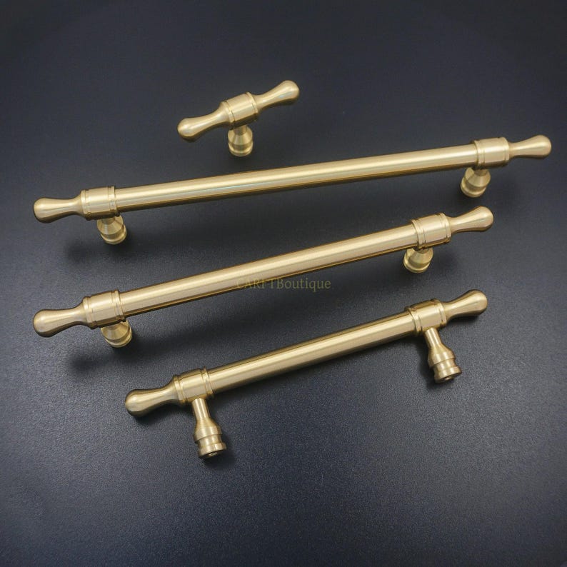K&ouml;nnte beinhalten: Ein Set aus vier messingfarbenen Schrankgriffen und -kn&ouml;pfen. Das Set enth&auml;lt drei lange Stangengriffe und einen kleineren Knopf. Die Hardware hat ein klassisches Design mit einem zylindrischen Stab und dekorativen Endst&uuml;cken. Auf dunklem Hintergrund.