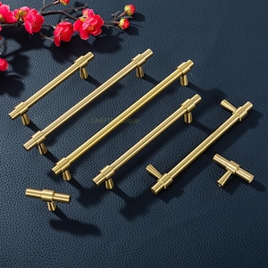 May include: A collection of gold-colored cabinet pulls and handles in various sizes. The pulls have a cylindrical bar design with a T-shaped base. The hardware is arranged on a dark blue surface, with a sprig of red flowers in the background.