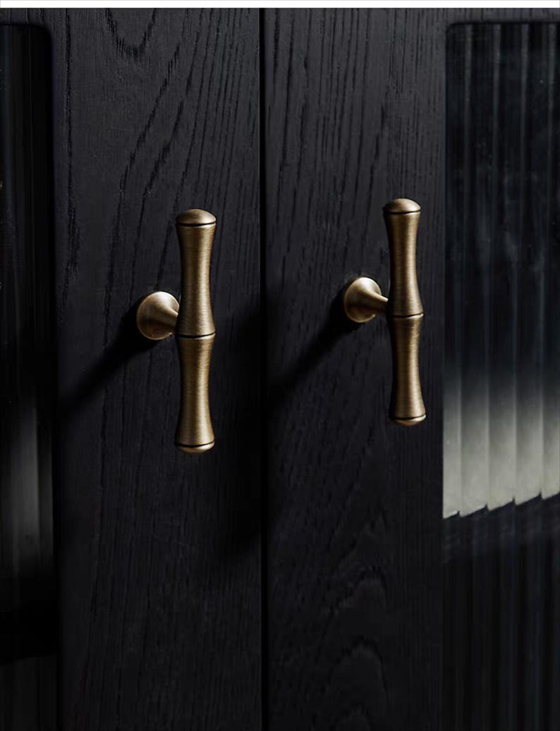 K&ouml;nnte beinhalten: Nahaufnahme einer schwarzen Holzschrankt&uuml;r mit zwei messingfarbenen Griffen im Bambus-Stil. Die Griffe sind vertikal ausgerichtet und haben eine zylindrische Form mit einer leichten Kr&uuml;mmung. Die Holzmaserung ist sichtbar.