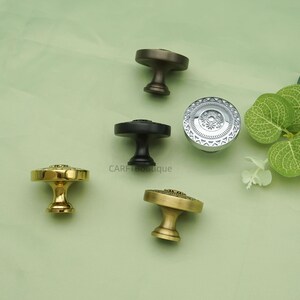 May include: An assortment of decorative cabinet knobs in various metallic finishes, including gold, silver, and bronze. Each knob features a unique design, with intricate patterns and textures. The knobs are arranged on a light green surface.