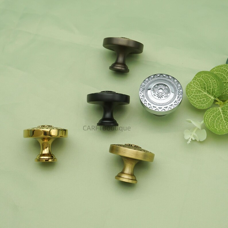 May include: An assortment of decorative cabinet knobs in various metallic finishes, including gold, silver, and bronze. Each knob features a unique design, with intricate patterns and textures. The knobs are arranged on a light green surface.