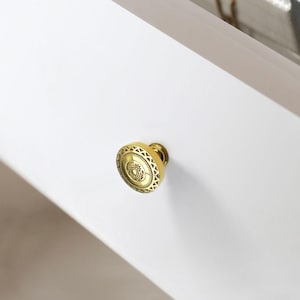 May include: A gold-colored cabinet knob with an ornate, circular design. The knob is mounted on a white surface, and the design features a central floral motif surrounded by geometric patterns. The hardware is likely for furniture or cabinetry.